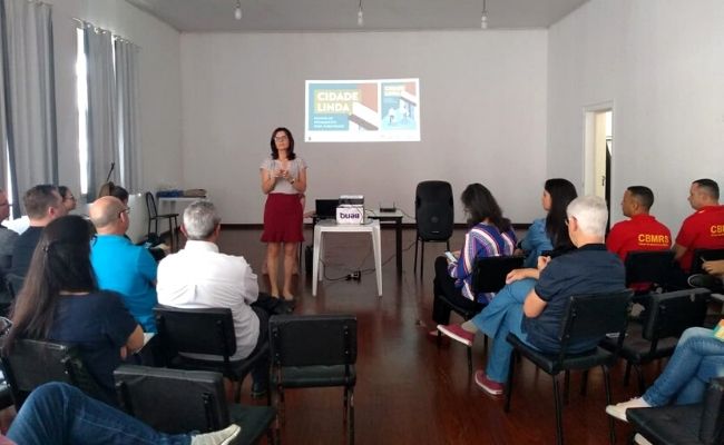 Em uma sala, em pé, ao fundo, a secretária Roberta e público sentado em cadeiras