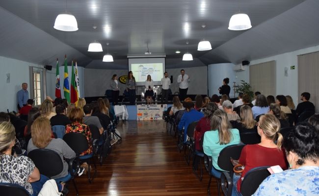Em um auditório, ao fundo, um palco com quatro pessoas em cima. Público sentado em cadeiras com um corredor no meio 