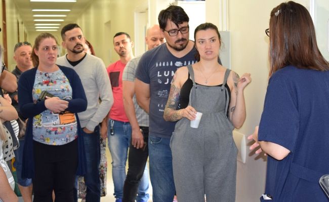No corredor da maternidade, uma médica em pé de costas e casais também em pé ouvindo orientações