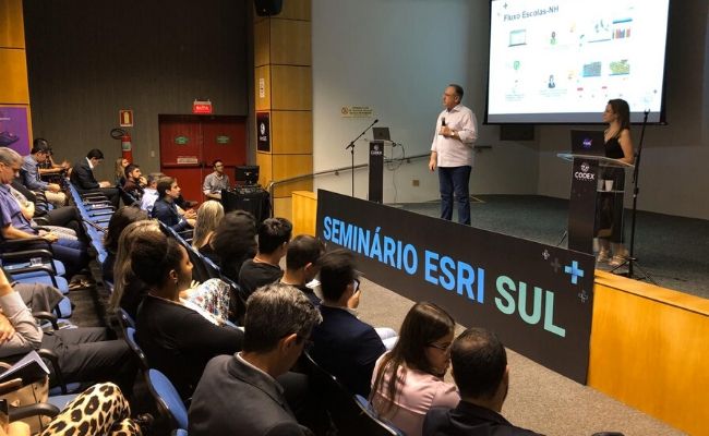 Em um palco, o secretário em pé ao microfone e público sentado a sua frente