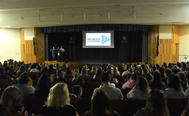 Ao fundo, palco com projeção de uma imagem. Público sentando em cadeiras