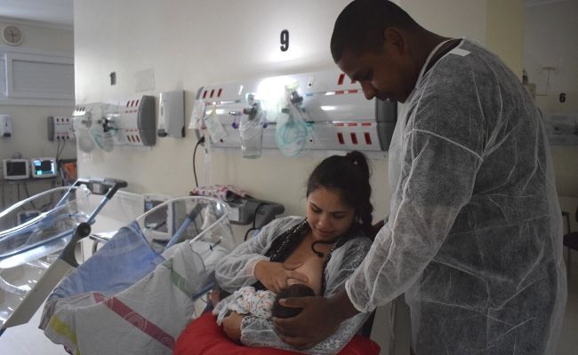 Na maternidade, mulher amamenta uma criança sentada e pai em pé ao lado