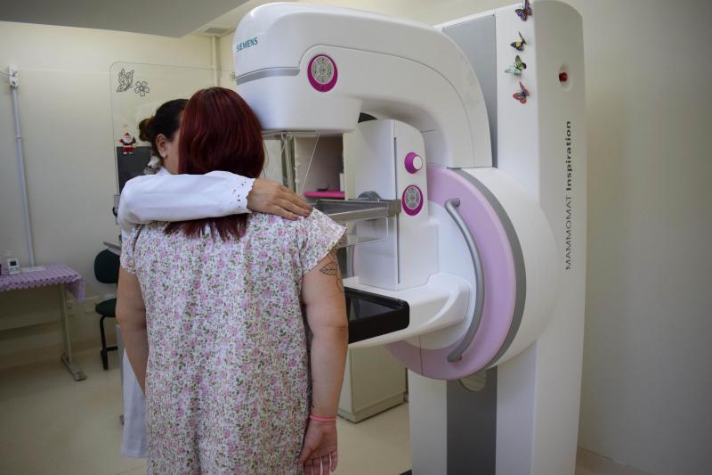Em uma sala com um mamográfo, mulher faz exame em pé no equipamento e uma enfermeira auxilia também em pé ao lado