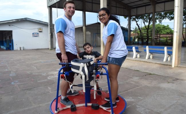 Em cima do brinquedo no átio da escola, que é em formato redondo, os produtores Marcos Vinícius e Ana Carolina e o aluno Marcos Vinícius