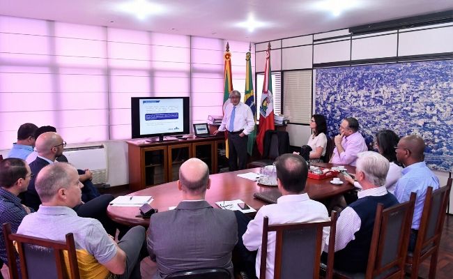 Em uma sala, homem em pé ao fundo ao lado de uma televisão, enquanto outras pessoas estão sentadas em semicírculo atrás de um mesa