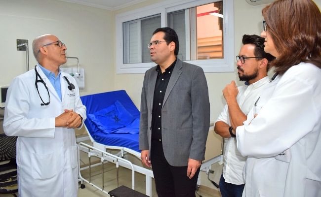 Em um quarto de hospital, com uma maca atrás, três homens e uma mulher em pé conversando