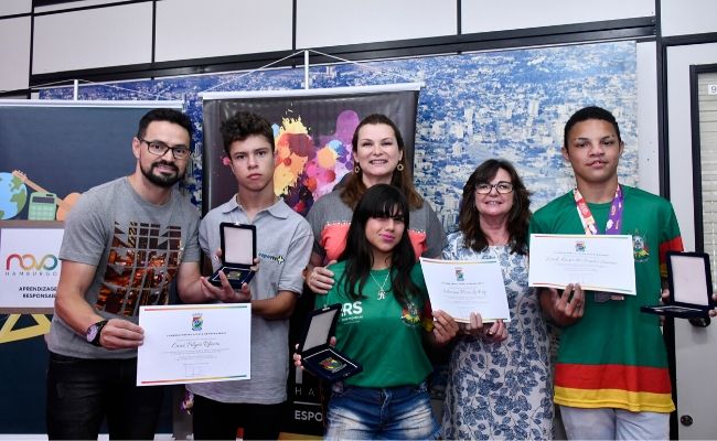 Em pé, lado a lado, a prefeita Fátima, o secretário Preto, a secretária Maristela e os três atles segurando o certificado