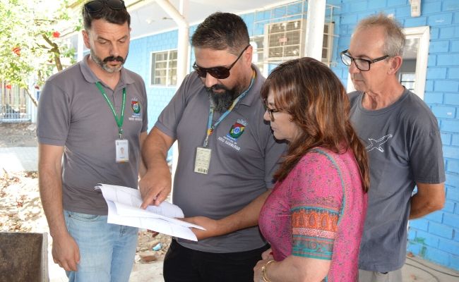No pátio de uma escola, três homens e a secretária Maristela em pé olham o projeto impresso em um papel
