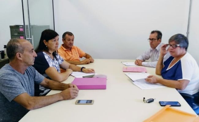 Em uma sala, com uma mesa no centro, três pessoas sentadas à esquerda, e duas à direita