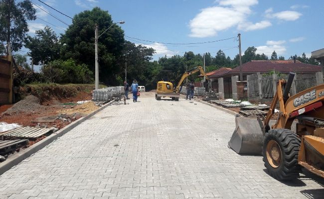 A imagem mostra uma rua pavimentada com blocos de concreto e uma máquina retroescavadeira estacionada à direita e homens trabalhando no fim da via