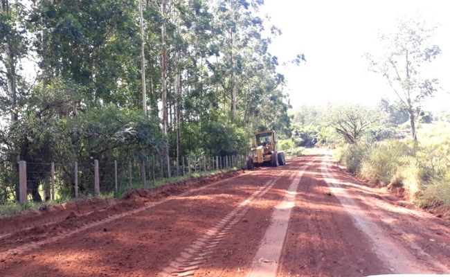 A imagem mostra uma estrada de terra e uma patrola fazendo o patrolamento