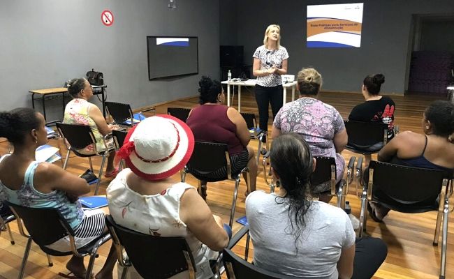 A imagem mostra uma sala, com alunas sentadas de costas em cadeiras e uma professora em pé ao fundo