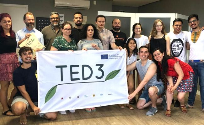 A imagem mostra um grupo de pessoas segurando um pano escrito TED 3