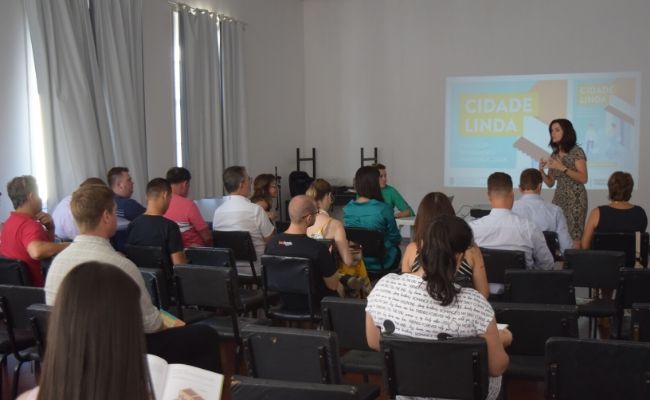 A imagem mostra uma sala com pessoas sentadas de costas em cadeiras e a secretária Roberta em pé apontando para a projeção de imagem que está na parede dos fundos da sala
