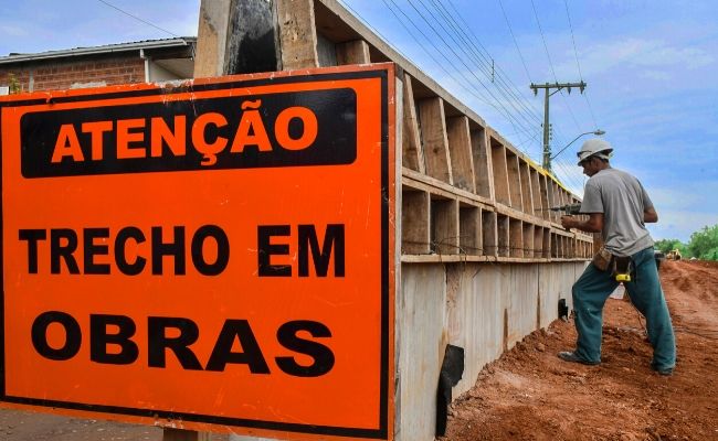 A imagem mostra uma placa laranja com letras pretas escrito "atenção trecho em obras" e um muro de concreto com um homem fazendo um furo com uma furadeira