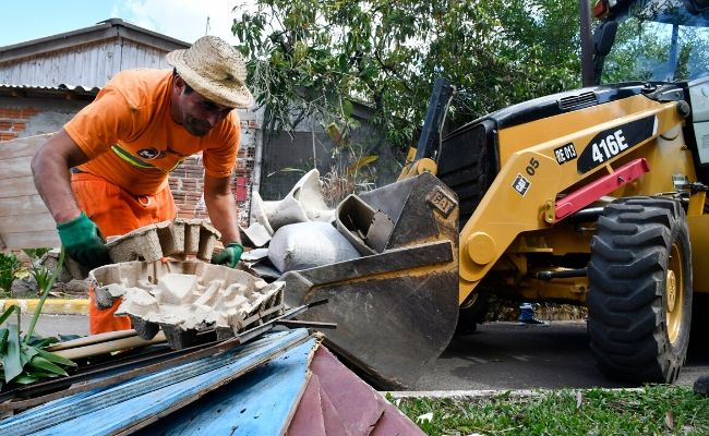 A imagem mostra uma retroescavadeira estaciona e um servidor vestindo laranja recolocando madeiras dentro da "concha" do equipamento