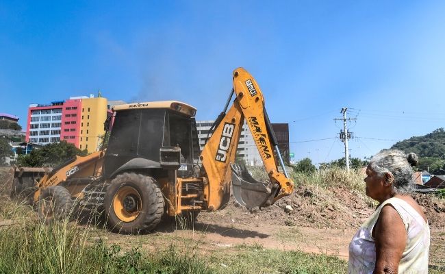 A imagem mostra uma senhora em pé olhando para uma retroescavadeira que patrola um terreno