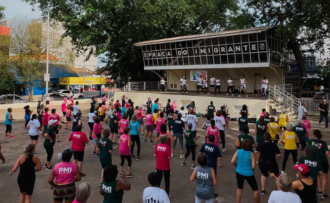 Mega aula de dança na Praça do Imigrante, na área central da cidade, com um grande número de participantes da melhor idade usando camisetas coloridas da PMI
