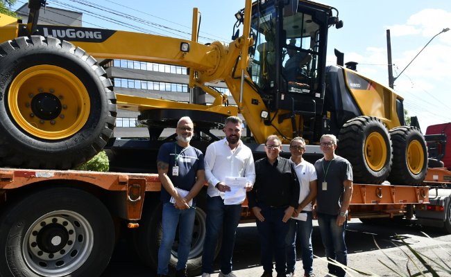 Prefeito Gustavo Finck ao lado do vice-prefeito Gerson Haas e demais equipe que compareceu à solenidade estão parados um do lado do outro em frente a uma motoniveladora amarela