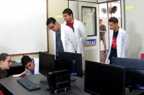 Em uma sala com computadores, quatro alunos em pé e de jalecos brancos, vistoriam a sala