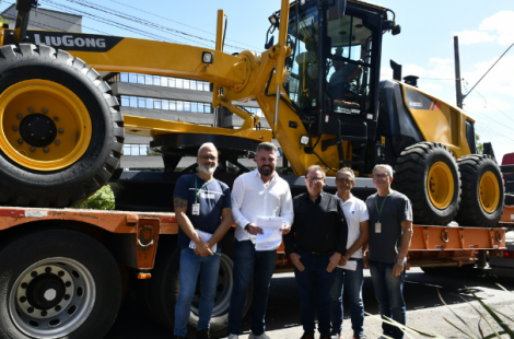Prefeito Gustavo Finck ao lado do vice-prefeito Gerson Haas e demais equipe que compareceu à solenidade estão parados um do lado do outro em frente a uma motoniveladora amarela